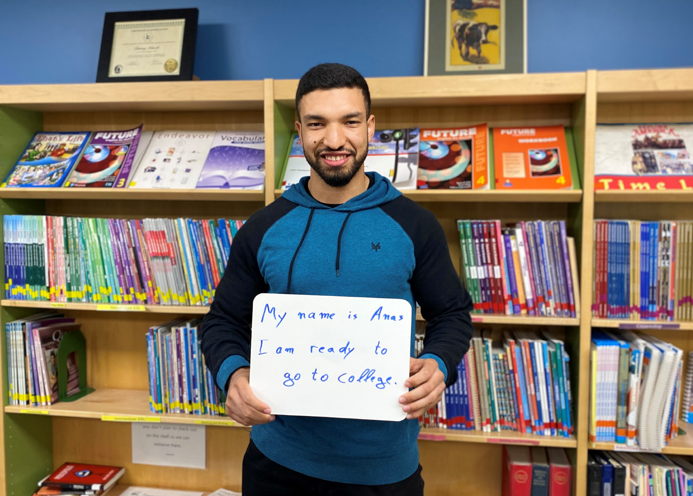 Man in a library holding a sign that says "My name is Ana and I am ready to go to college."