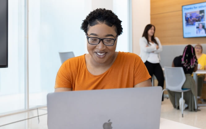 A woman in the Ascendium break room smiles down at a laptop while her colleagues converse in the background