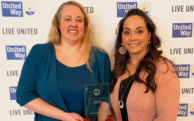 Two women from Ascendium stand smiling in front a United Way wall logos holding a glass award